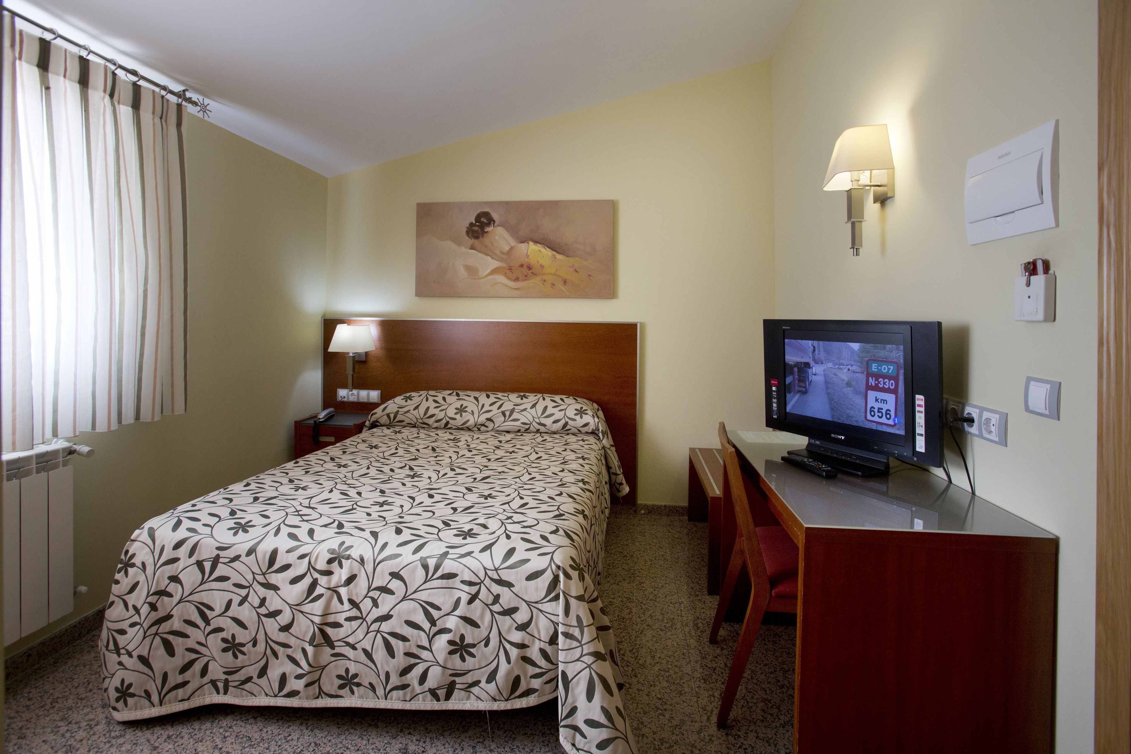 A hotel room with a bed, a desk with a television, a painting on the wall, and a window with curtains.