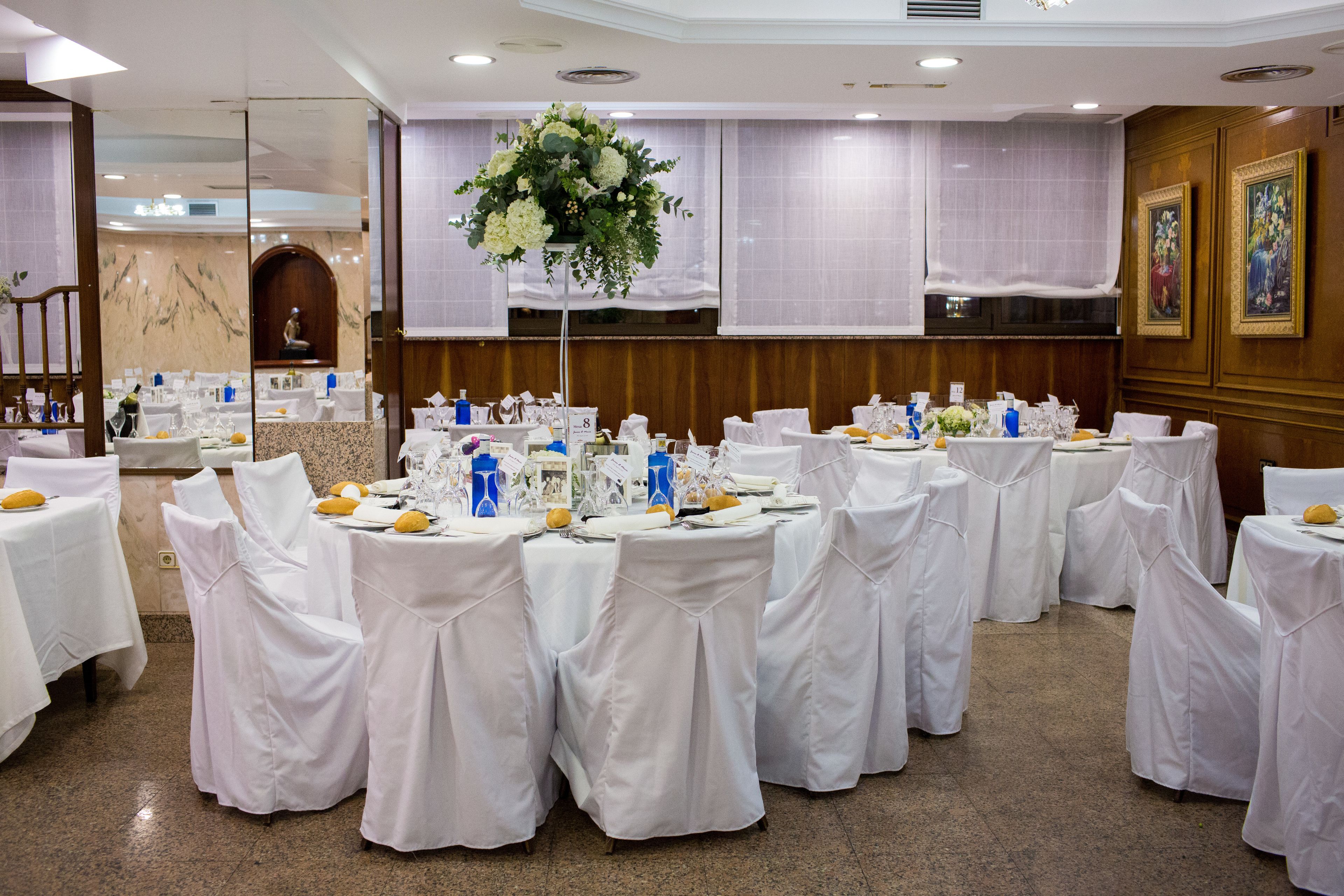 Una sala de banquetes amb taules rodones cobertes amb teles blanques, cadires amb fundes blanques i un arranjament floral central.