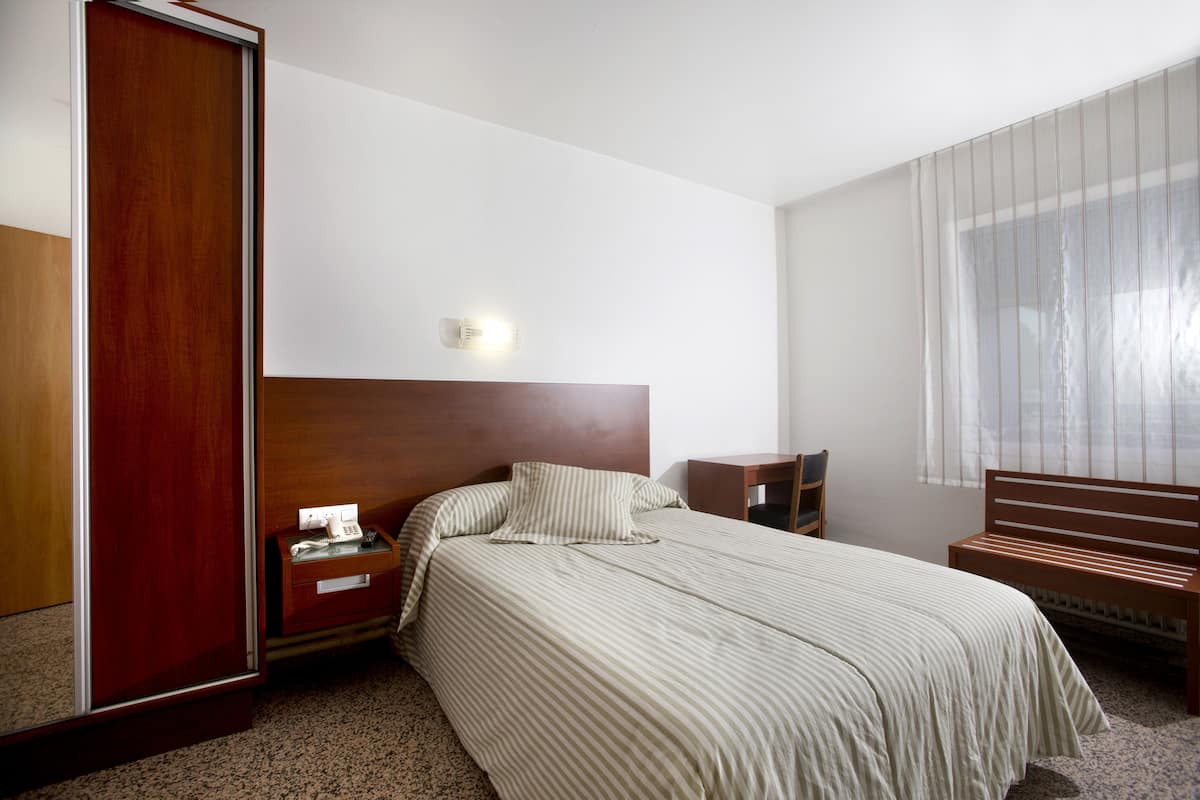 A hotel room with a large bed, a wooden headboard, a nightstand, a bench, and a window with sheer curtains.