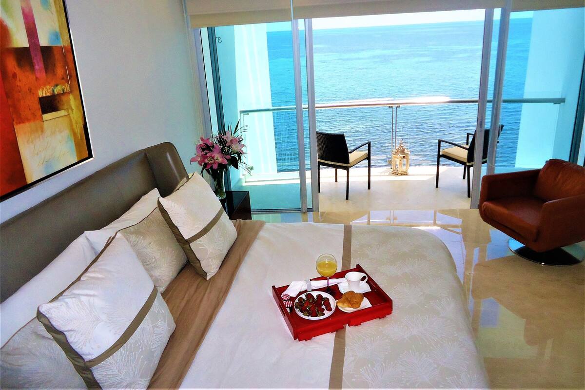 A hotel room with a bed, a breakfast tray, a vase with flowers, and a view of the ocean.