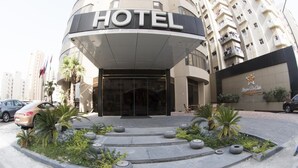 A hotel entrance with a curved facade, a glass door, and a landscaped area with palm trees and planters. - Lumiere Des Etoile (Salmiya)