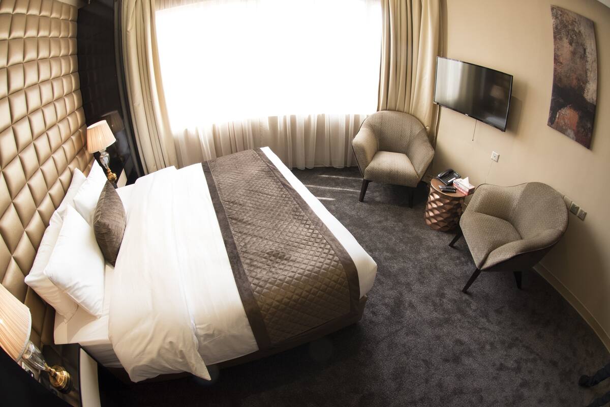 Hotellihuone, jossa on sänky, kaksi nojatuolia, televisio ja suuri ikkuna, jossa on verhot.