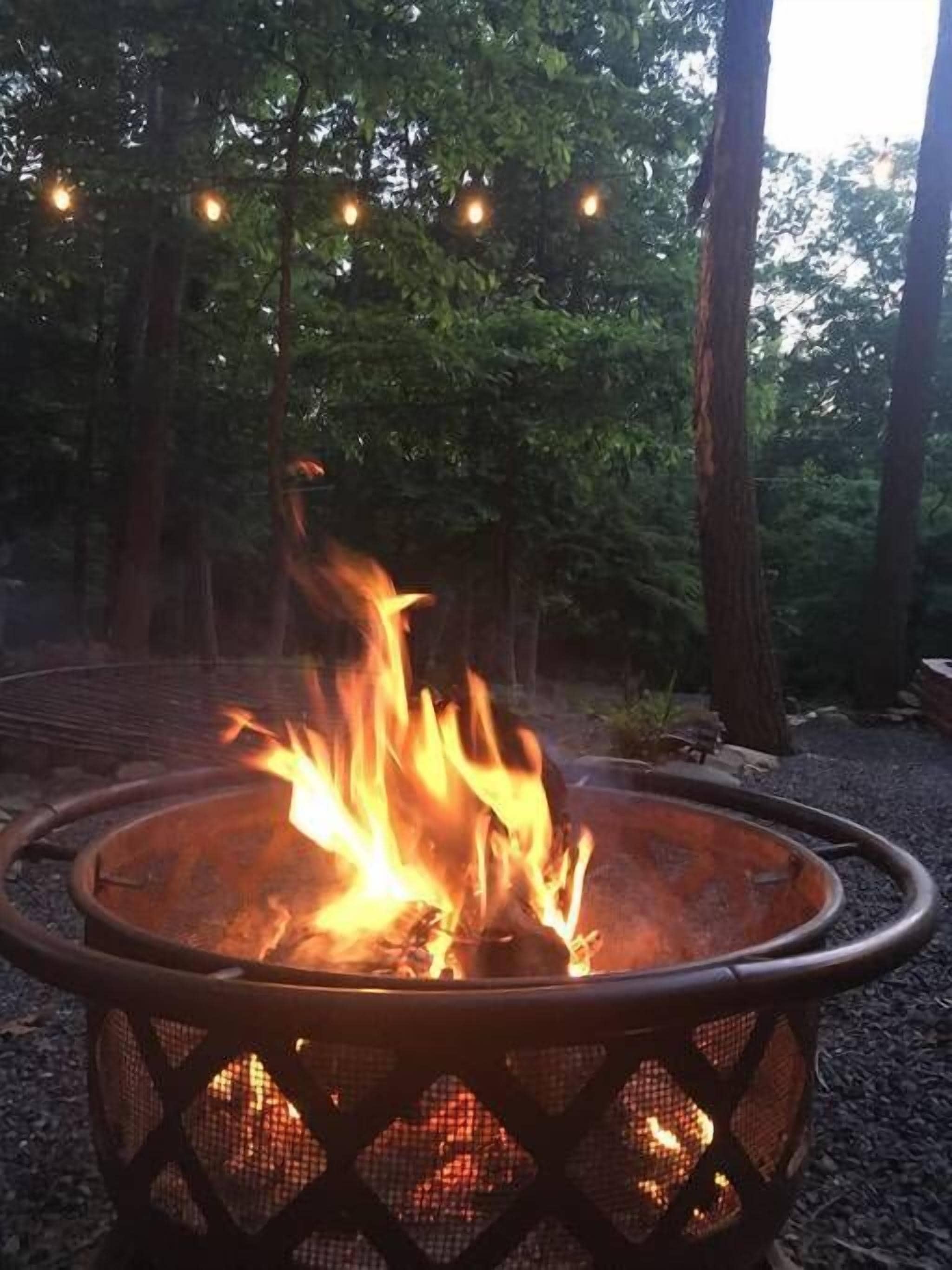 A fire pit with flames, surrounded by trees and string lights.