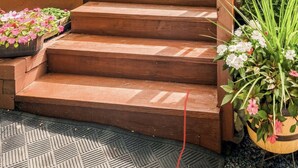 Eine Holzterrasse führt zu einem Eingang mit Glastür, umgeben von Grünpflanzen und Gartenmöbeln.
