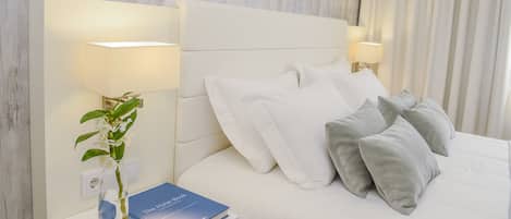 A neatly made bed with white and gray pillows, a bedside table with a lamp, a vase with flowers, and a book titled "Hotel Book".