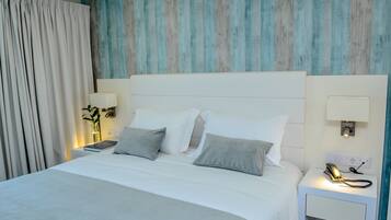 Una cama bien hecha con ropa de cama blanca y gris, una lámpara de noche y un teléfono sobre una mesita de noche.