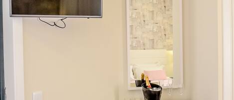 A hotel room with a TV mounted on the wall, a small table with a bottle of champagne, a chair, and a mirror with a decorative frame.