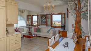 Een woonkamer met een witte bank, een houten salontafel en een eettafel met stoelen. Er is een kroonluchter, een groot muurschildering van een boom en uitzicht door de ramen.