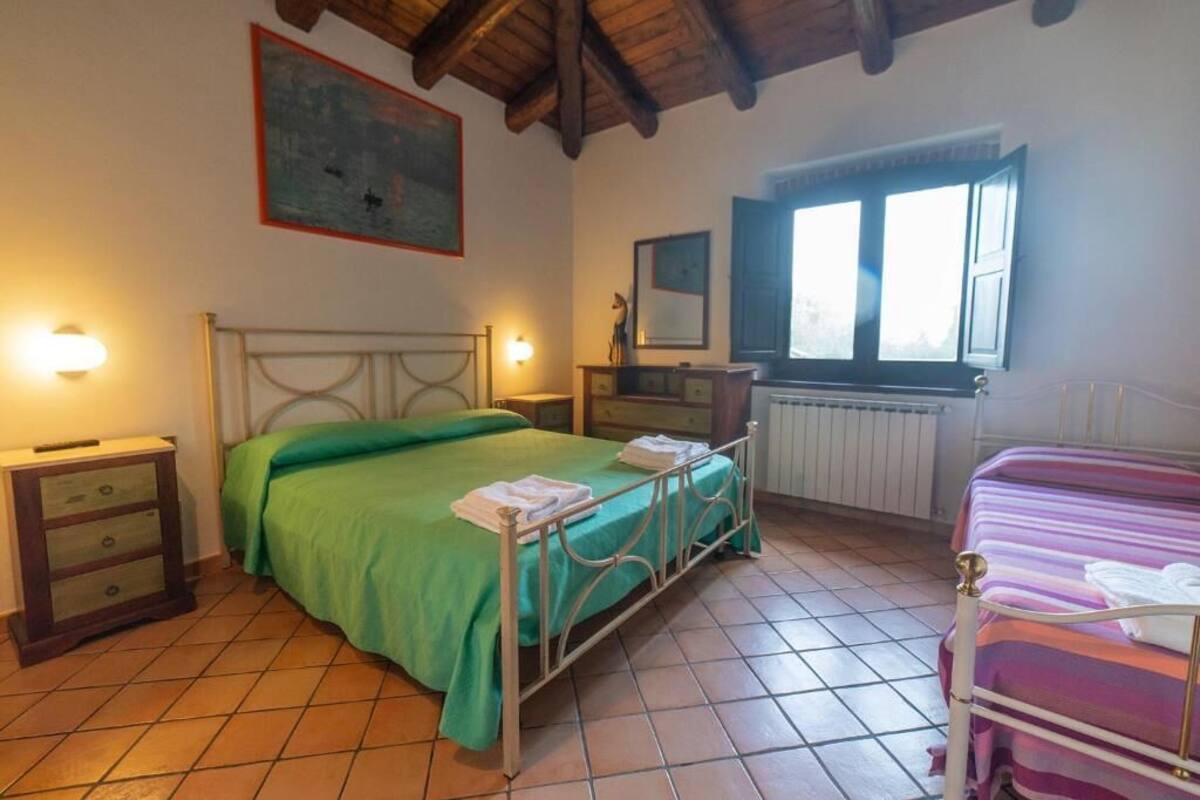 Une chambre avec un lit en métal, une literie verte, une commode en bois, une fenêtre avec des volets ouverts et un radiateur.