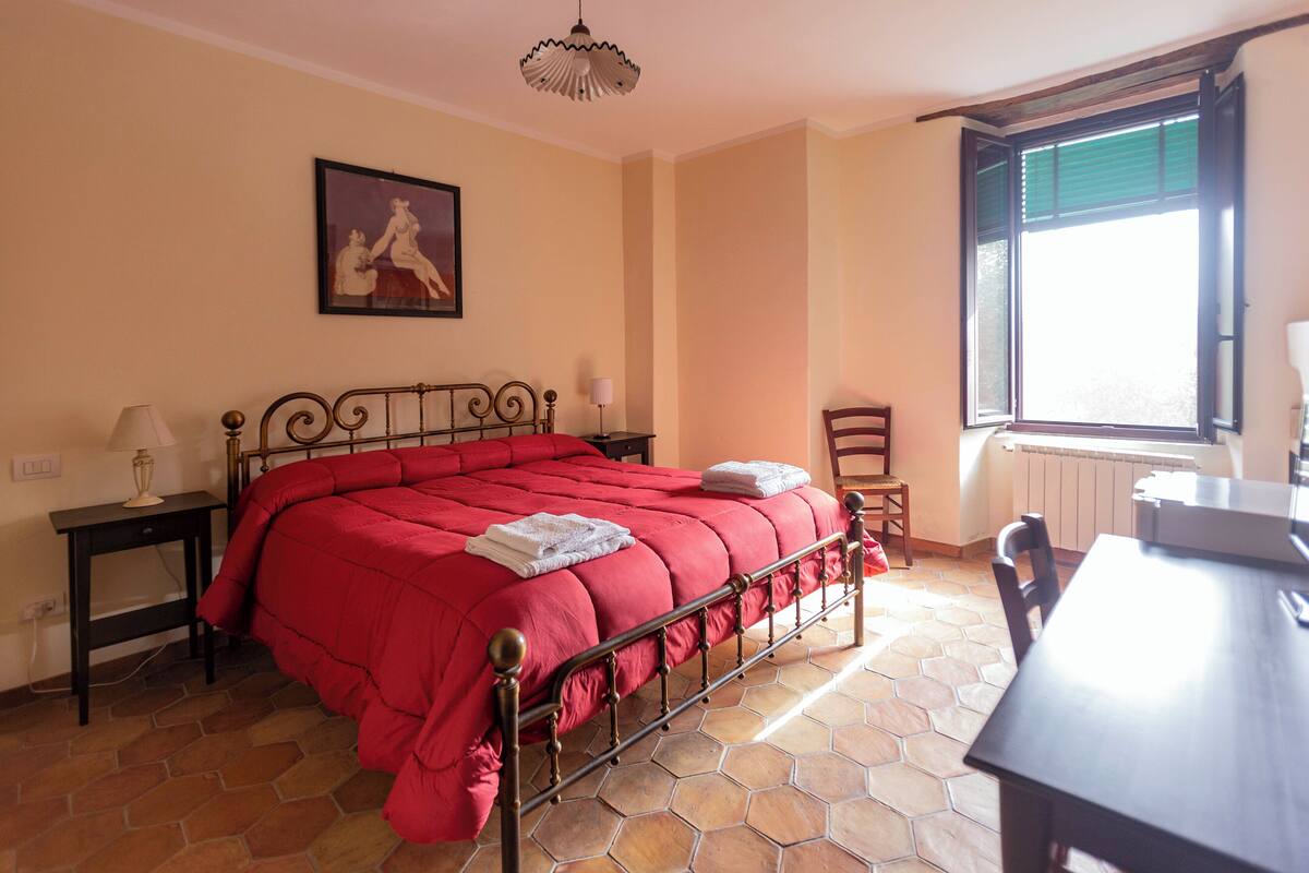 Une chambre à coucher avec un lit, une chaise, une table, une lampe, un tableau au mur et une fenêtre.