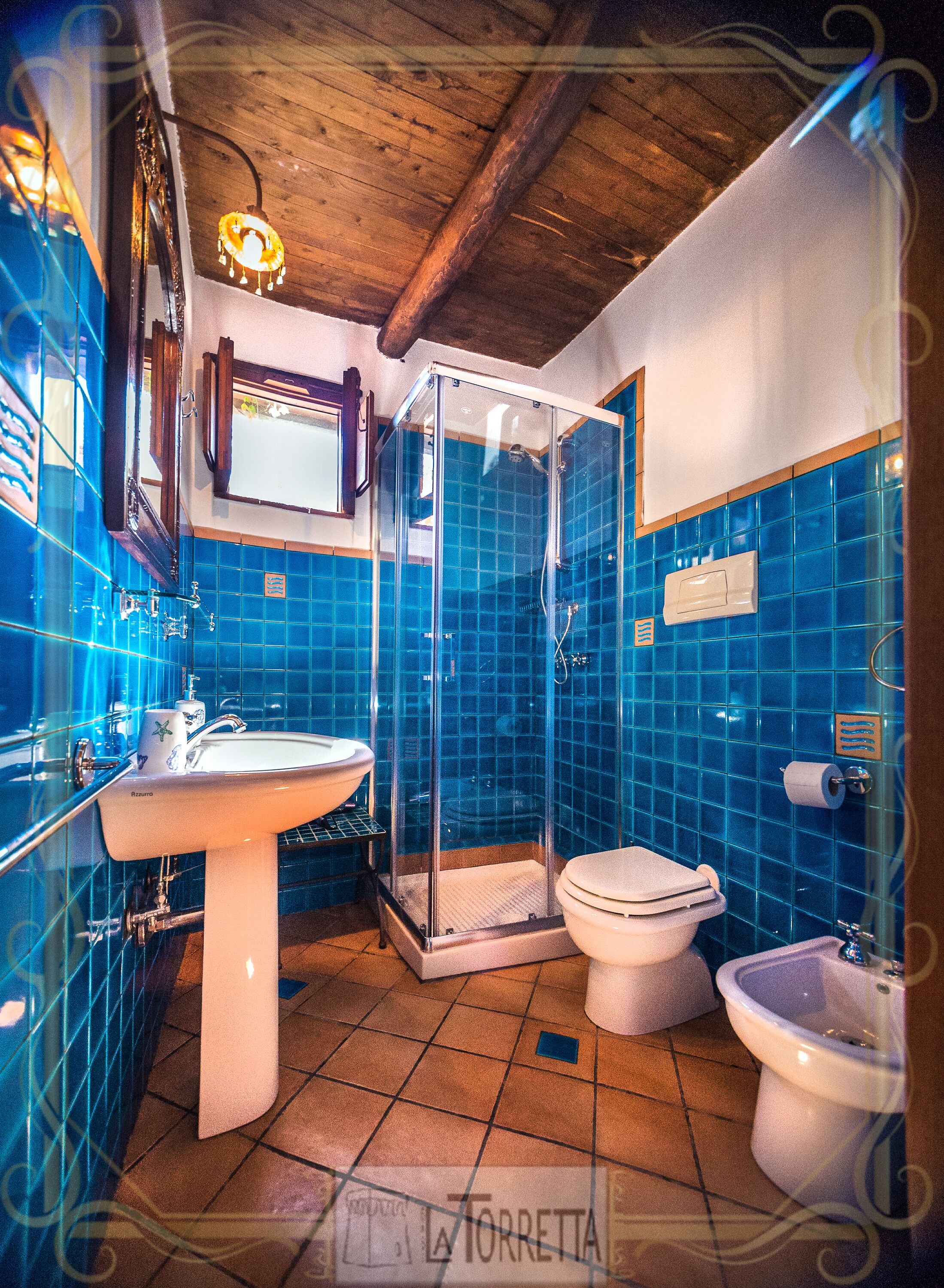 Ein Badezimmer mit einer gläsernen Duschkabine, einem Waschbecken, zwei Toiletten und blautonierten Fliesen.