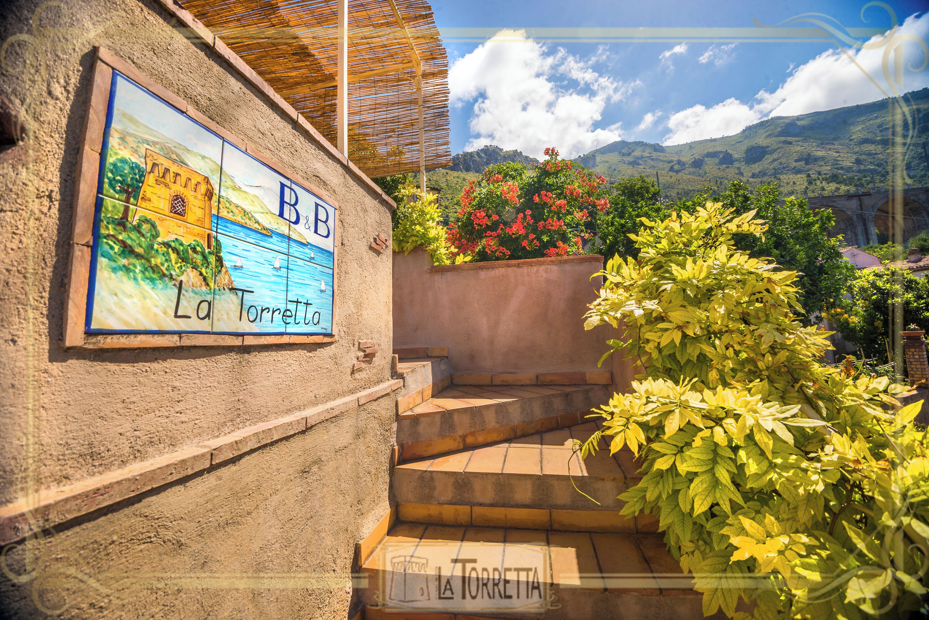 A building with a sign reading 'B&B La Torretta' and a staircase leading to a terrace with plants and a view of hills.