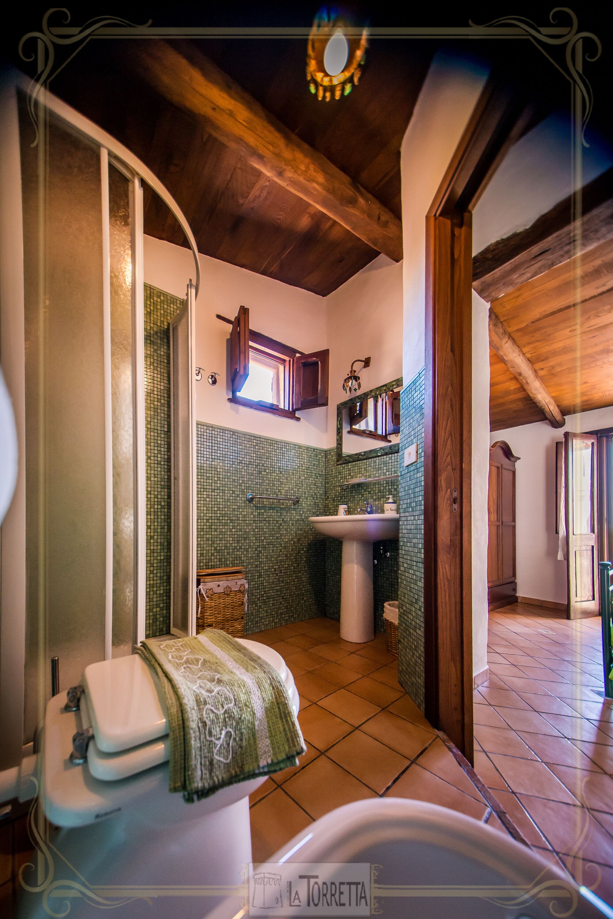 Baño con inodoro, lavamanos y zona de ducha, con azulejos verdes y vigas de madera.