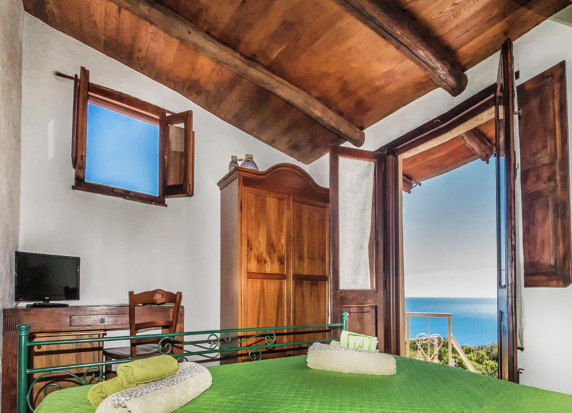 A bedroom with a bed, a wooden wardrobe, a television, and a view of the sea.