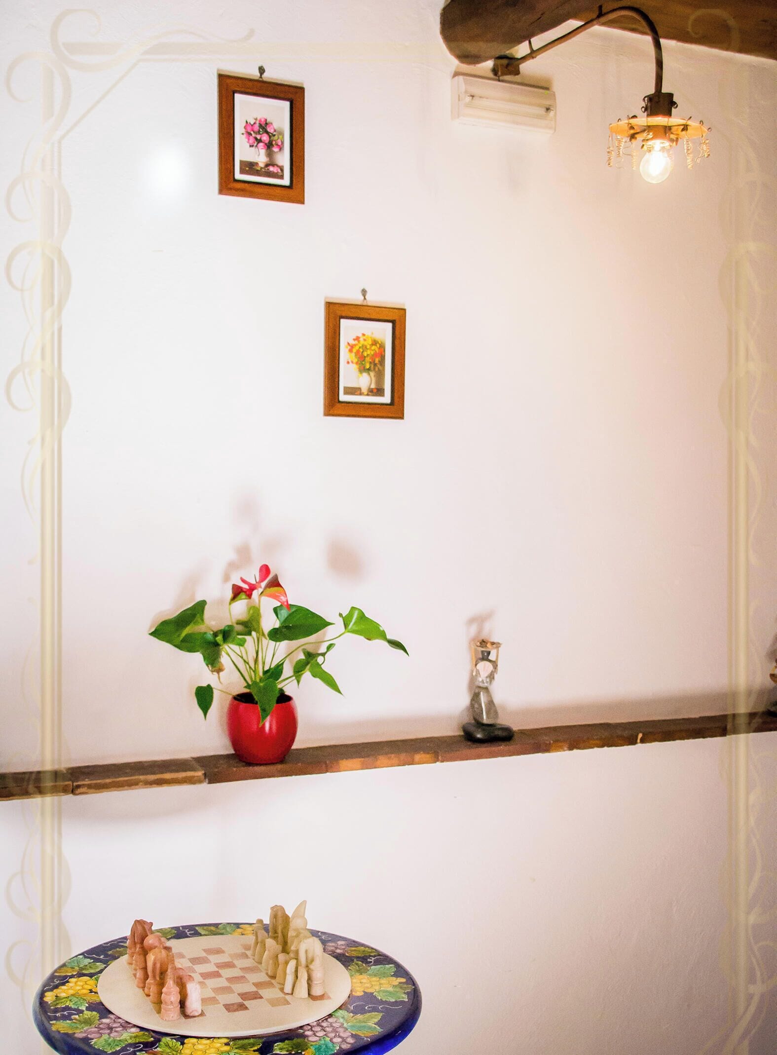 A room with a white wall, a wooden shelf, two framed pictures, a potted plant, a small figurine, and a chessboard.
