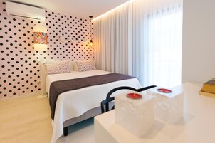 Una habitación de hotel moderna con una pared de lunares, una cama con colcha blanca y marrón, una mesita de noche con una vela y un ventanal con cortinas.