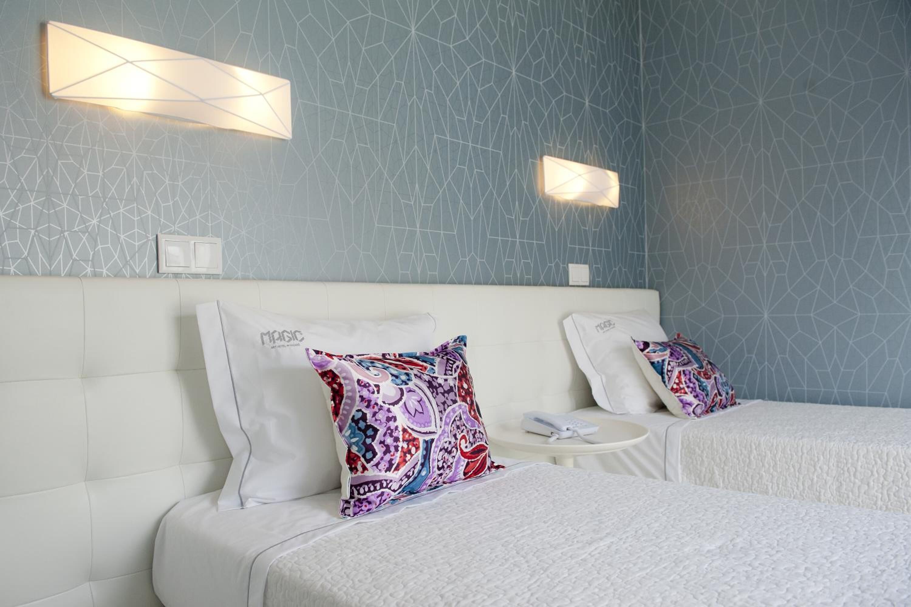 A hotel room with two beds, a white headboard, patterned wall, and two wall-mounted lights.