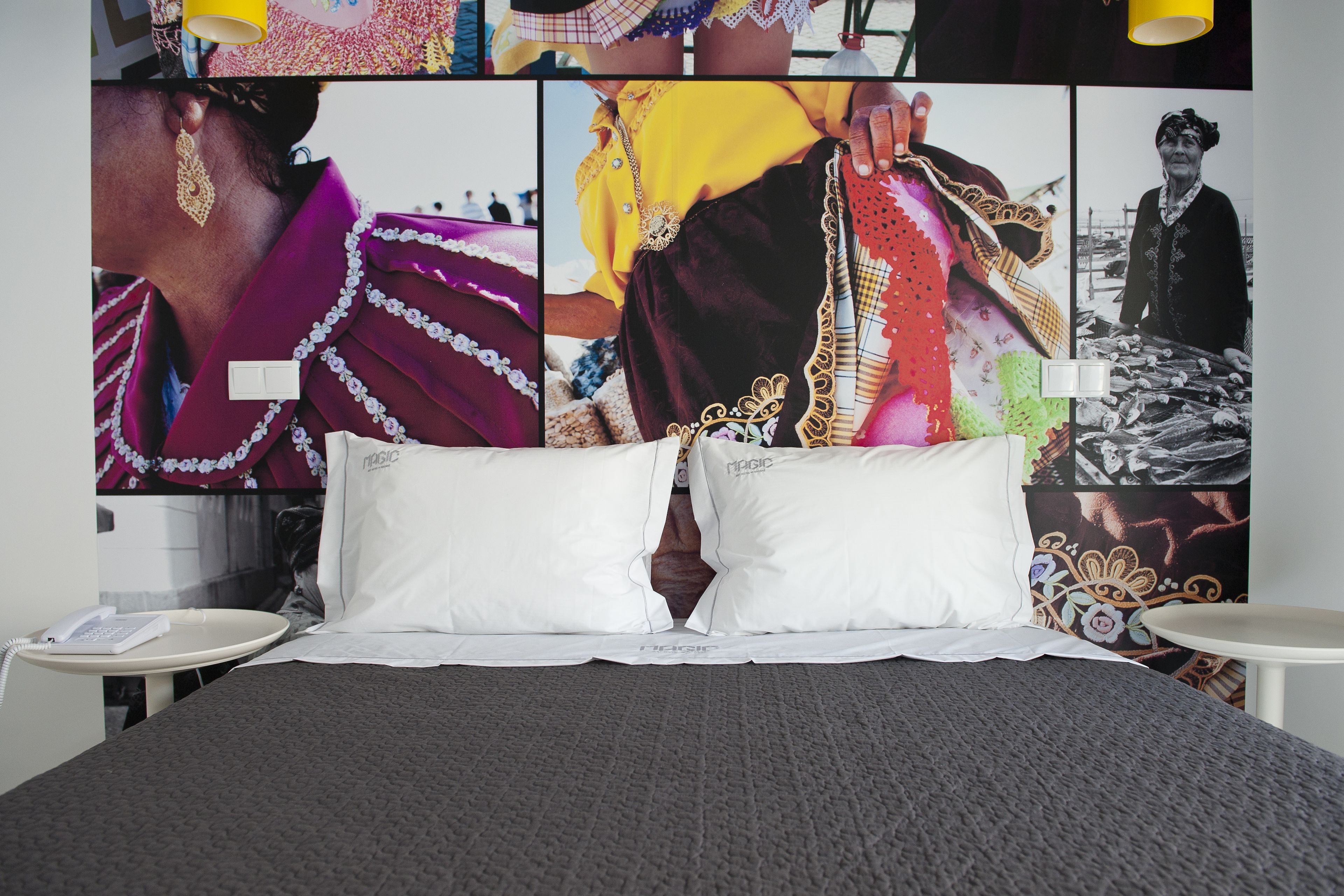 A hotel room with a large bed, white pillows, a grey blanket, and a wall decorated with colorful photographs.