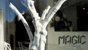 Une sculpture blanche et abstraite, placée dans un pot de fleurs blanc à motifs géométriques, se trouve devant un bâtiment à la façade en verre, sur lequel on peut lire l'inscription « MAGIC ART HOTEL NAZARE ».