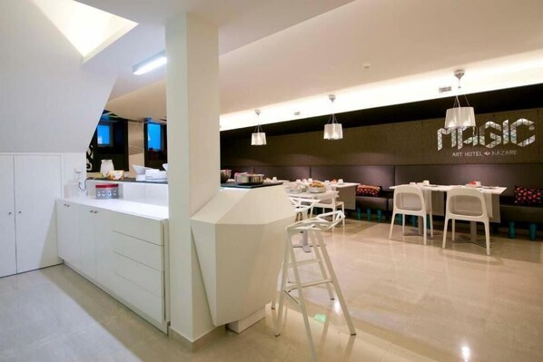A modern hotel lobby with a central island, white furniture, and a 'MAGIC ART HOTEL NAZARE' sign.