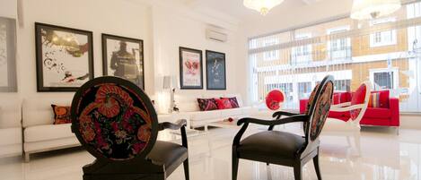A spacious living room with a white sofa, a red sofa, and two chairs with ornate backs. There are framed artworks on the walls and a chandelier hanging from the ceiling.
