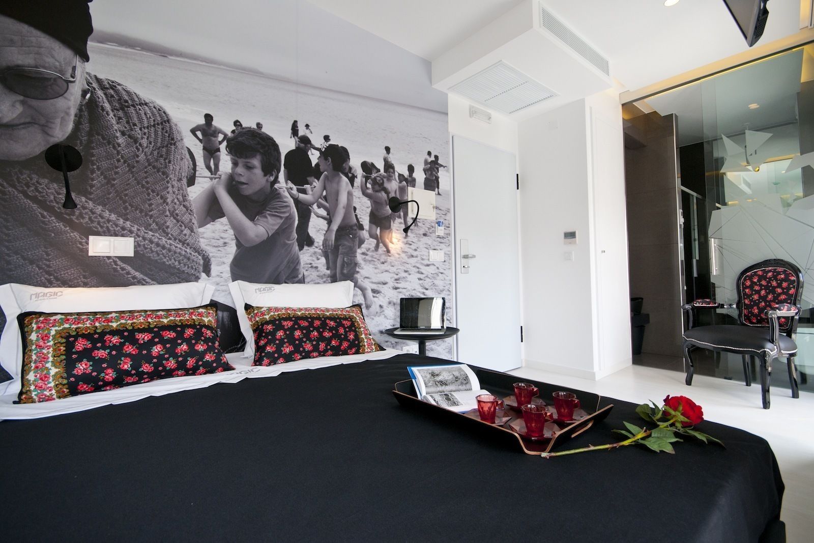A modern hotel room with a large bed, a glass partition wall, and a view of the beach.
