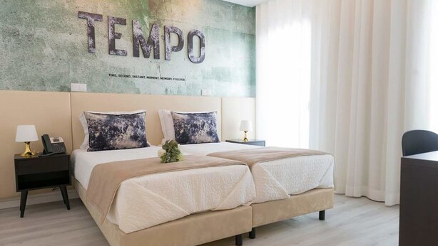Une chambre d’hôtel avec un grand lit, deux tables de chevet, une petite table et un mur sur lequel figure le mot « TEMPO » ainsi qu’un slogan.
