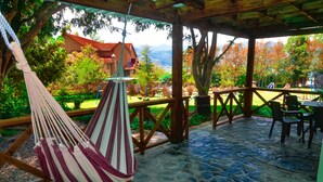 A covered outdoor area with a hammock, stone flooring, wooden furniture, and a view of a house and trees.