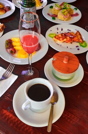 A dining table with plates of food, a glass of red drink, a cup of black coffee, and a dessert with a wooden lid.