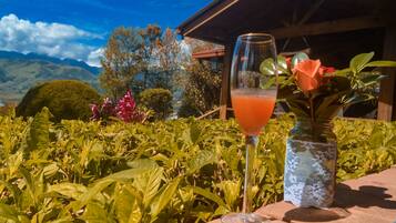 A glass of orange juice on a table with a lace-covered vase and a rose, set against a backdrop of lush greenery and mountains.