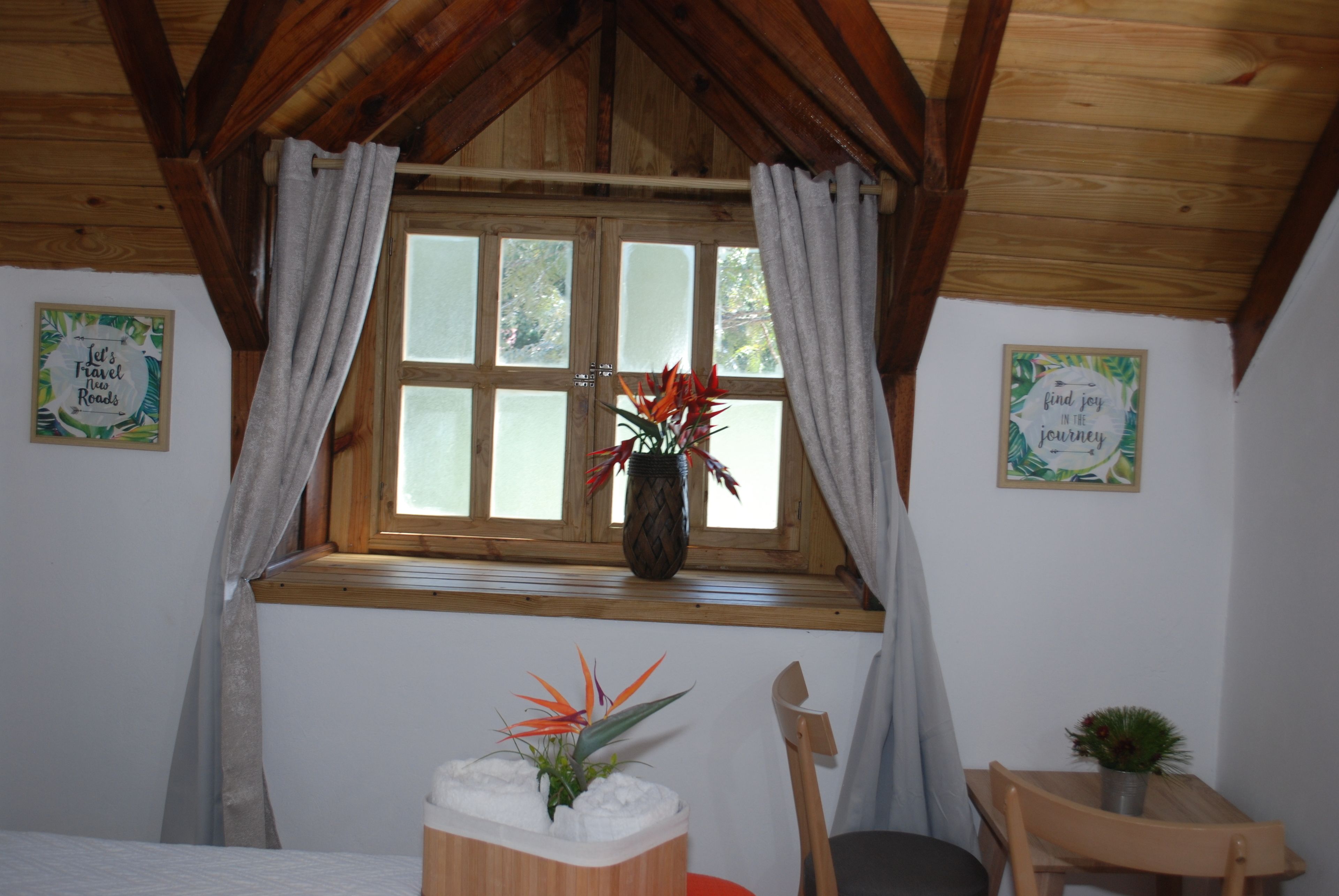 Une pièce chaleureuse avec un plafond en bois, une fenêtre avec des rideaux, un lit, une chaise et une petite table sur laquelle est posée une plante.
