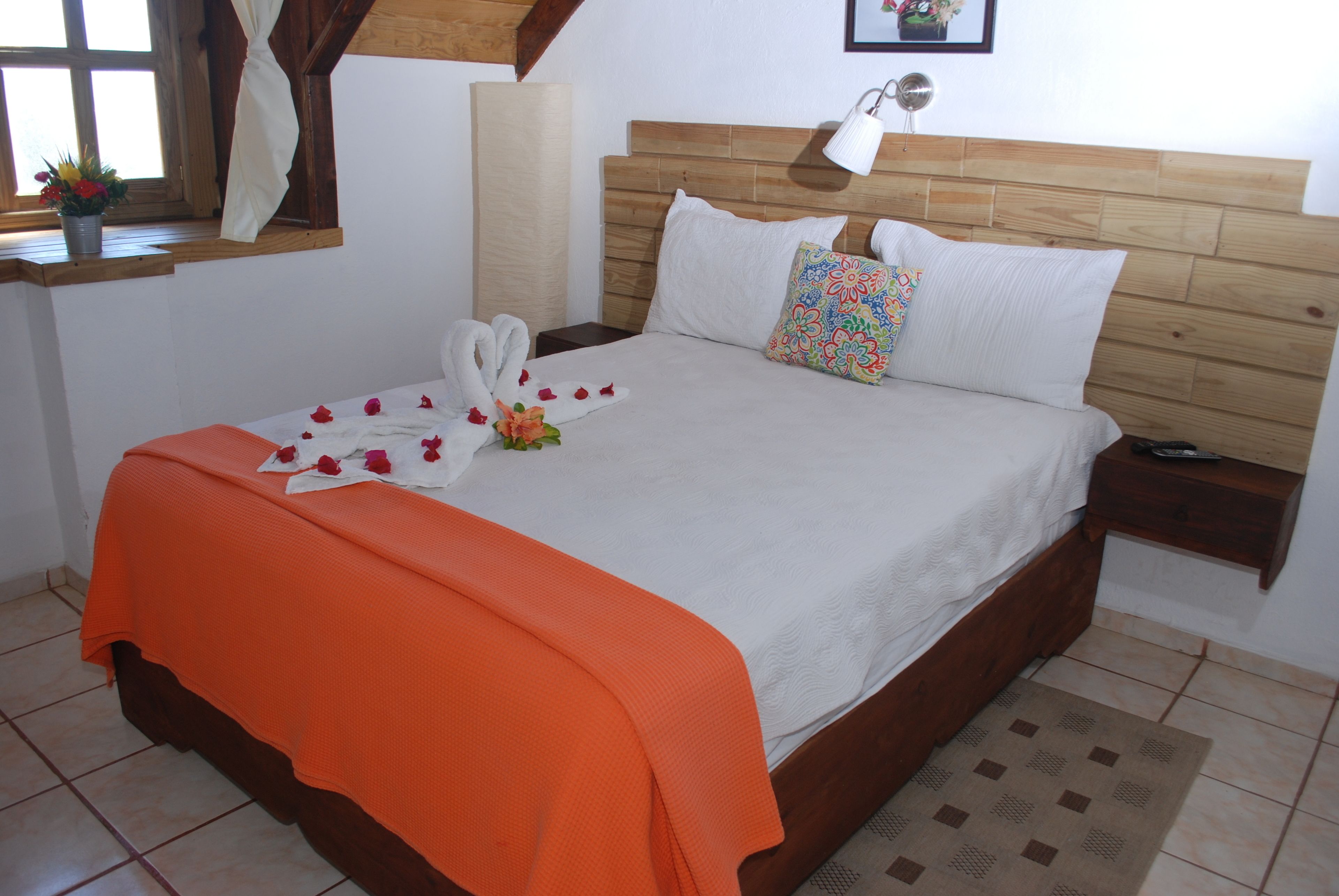 Una cama bien hecha con un edredón naranja, almohadas blancas y una almohada de colores. Hay una mesita de noche con una lámpara, una ventana con cortinas y una planta pequeña en el alféizar.