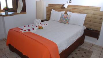 Una cama bien hecha con un edredón naranja, almohadas blancas y una almohada de colores. Hay una mesita de noche con una lámpara, una ventana con cortinas y una planta pequeña en el alféizar.