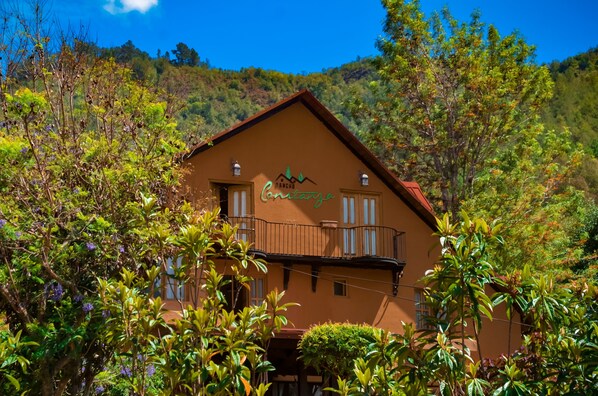 A two-story building with a balcony, surrounded by lush greenery and hills in the background.