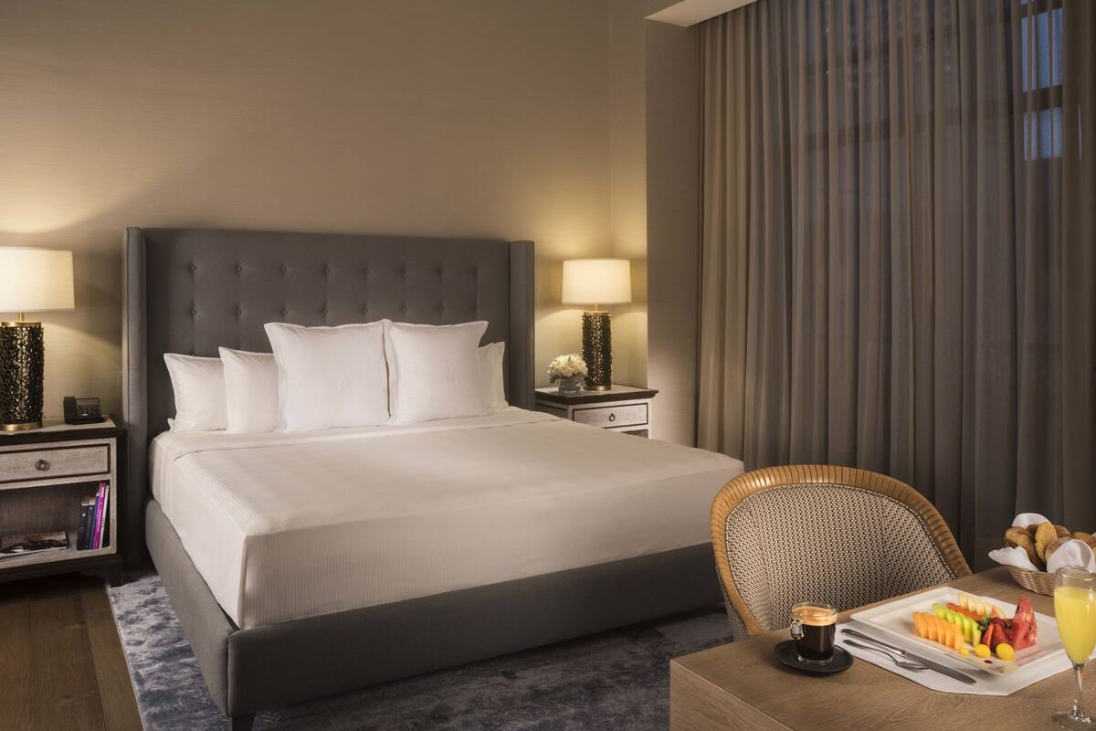 Une chambre d’hôtel avec un grand lit, deux tables de chevet avec des lampes, un fauteuil en osier, un plateau de petit-déjeuner avec des fruits et une tasse de café, et une vue sur la ville par la fenêtre.