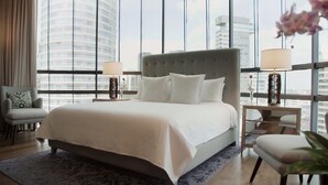 Quarto de hotel moderno com uma cama grande, duas mesas de cabeceira com luminárias, uma poltrona e vista para o horizonte da cidade através de amplas janelas.