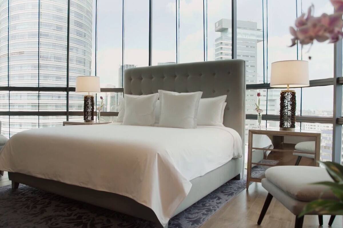 Une chambre d’hôtel moderne avec un grand lit, deux tables de chevet avec des lampes, un fauteuil et une vue sur le panorama de la ville, visible à travers de grandes fenêtres.