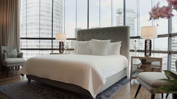 Une chambre d’hôtel moderne avec un grand lit, deux tables de chevet avec des lampes, un fauteuil et une vue sur le panorama de la ville, visible à travers de grandes fenêtres.