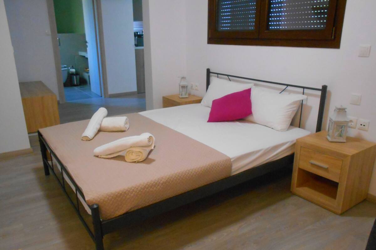 Una cama bien tendida con toallas enrolladas, una mesita de noche de madera con un cajón y una ventana con persianas.