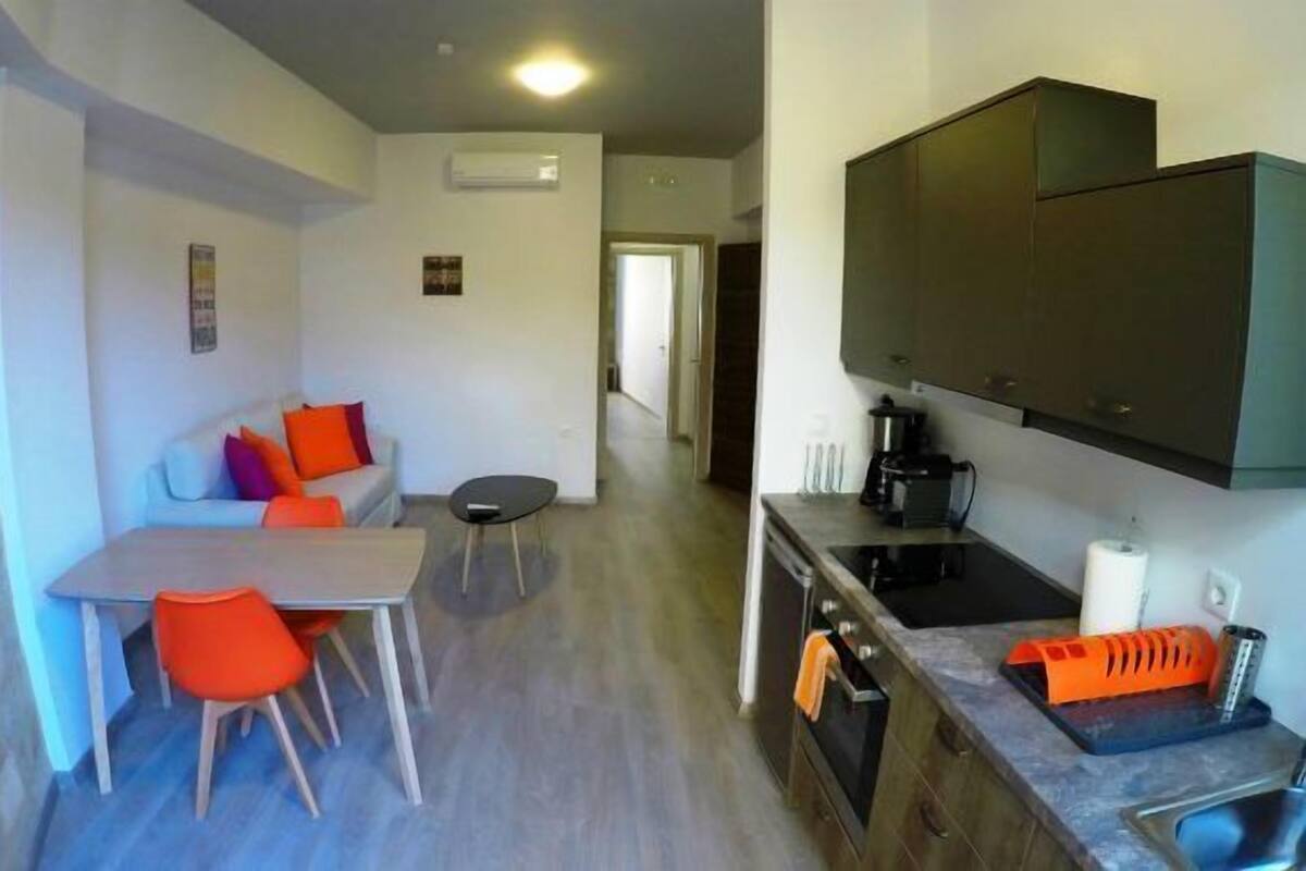 Un espacio habitable compacto con kitchenette, una zona de comedor con mesa y sillas, y un sofá con cojines de colores.