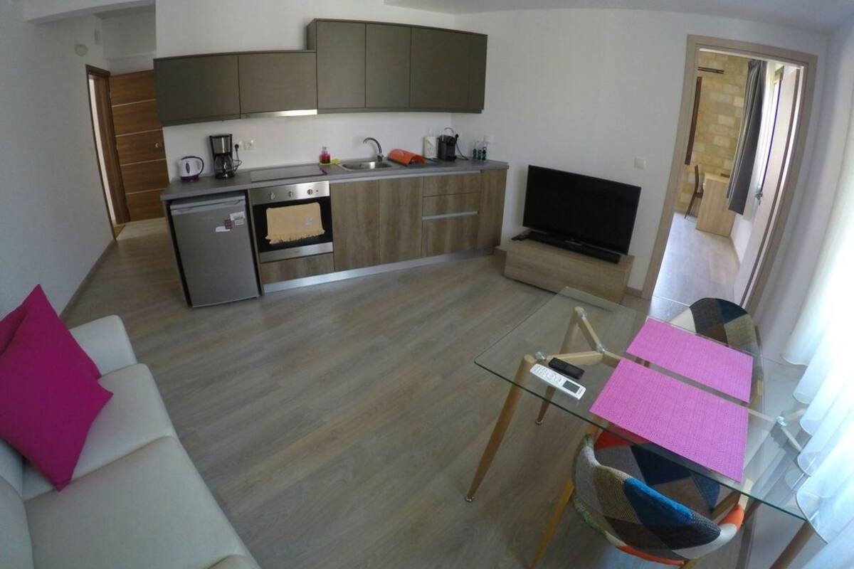 Una cocina moderna con horno empotrado, cafetera y televisión. Hay una mesa de comedor de vidrio con sillas.