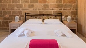Un lit bien fait, avec du linge de lit blanc et une serviette rose, deux oreillers blancs et une tête de lit en métal. Il y a deux tables de chevet avec des lampes blanches et des vases décoratifs. Le mur du fond de la pièce est en pierre.