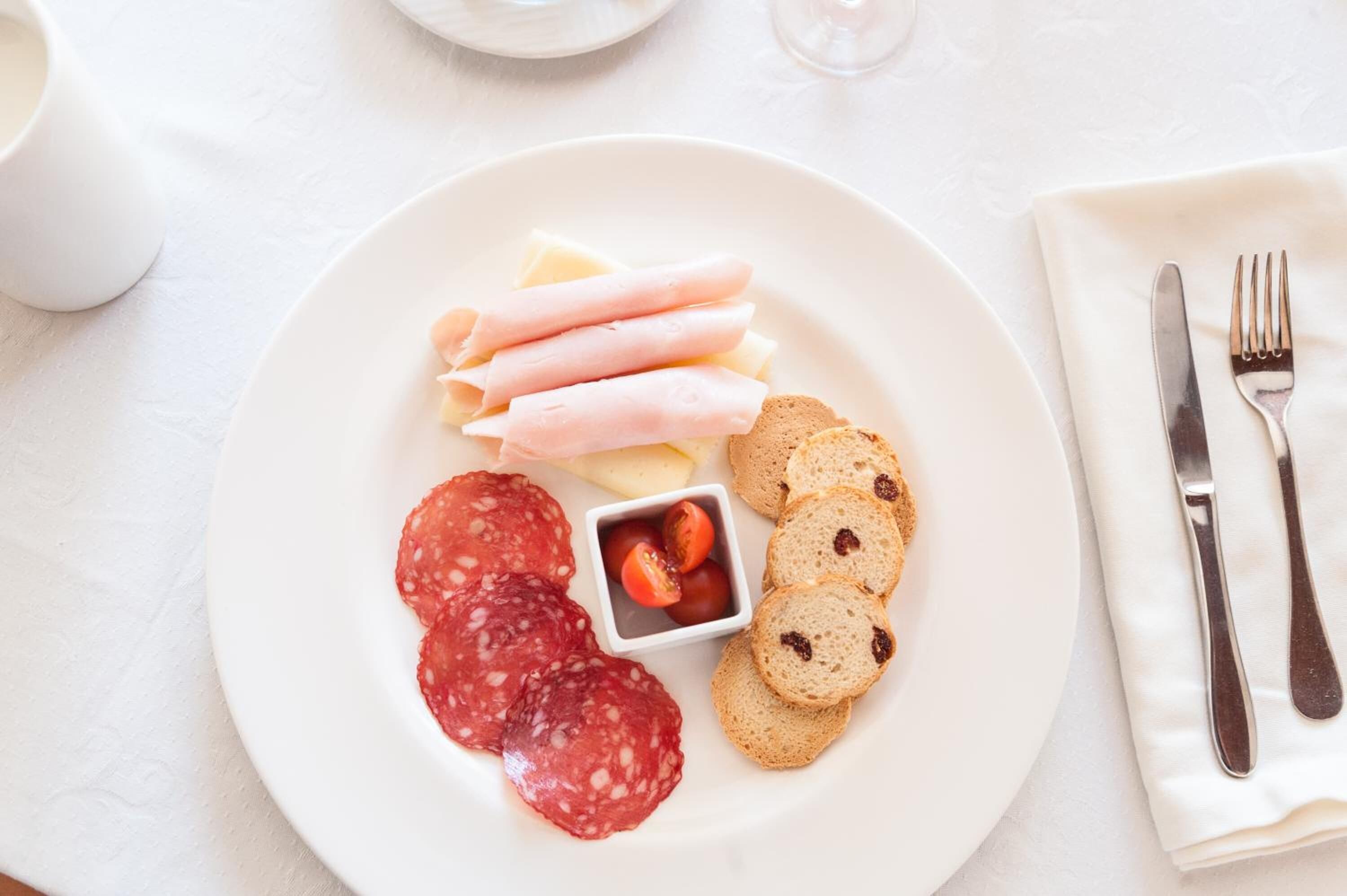 Une assiette blanche garnie de tranches de salami, de jambon, de fromage et de pain, le tout posé sur une nappe blanche.