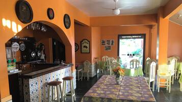 Um restaurante com uma mesa comprida, cadeiras, área de bar e vista através de uma porta.