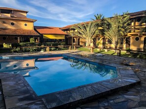 A swimming pool surrounded by stone tiles, with buildings featuring terracotta roofs and palm trees in the background. - Pousada Estalage (Cabo Frio)