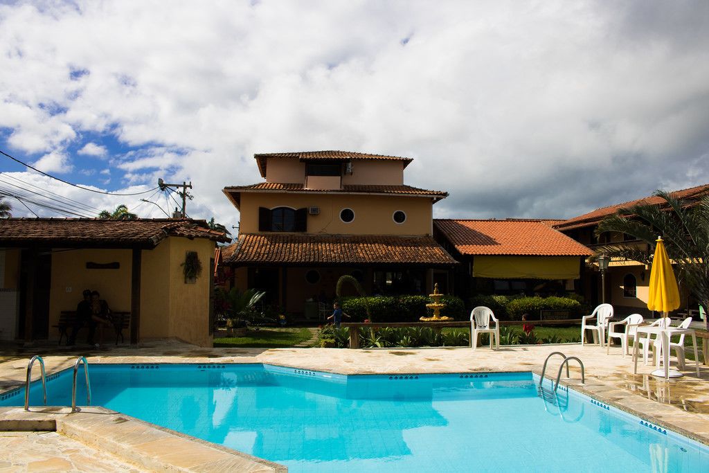 A swimming pool with a stone deck, surrounded by a two-story building with a tiled roof and a fountain.