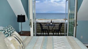 A bedroom with a bed, a nightstand, a lamp, a table with two chairs, and a view of the sea through an open door. - Stunning beach views on Over the Dunes - White Horse Beach, Plymouth (Plymouth)