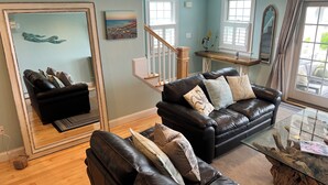 A living room with a black leather sofa, a glass coffee table, and a large mirror. - Stunning beach views on Over the Dunes - White Horse Beach, Plymouth (Plymouth)