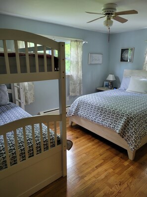A bedroom with a bunk bed, a single bed, a ceiling fan, a nightstand with a lamp, and a framed picture on the wall.