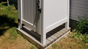 Een wit metalen hek met een zwarte hendel, bevestigd aan een grijs huis met een kleine veranda en een raam boven het hek.